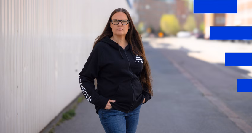 Pitkähiuksinen henkilö seisoo kaupunkikadulla katsoen vakavasti kameraan. Hänellä on musta huppari, jossa lukee "A-klinikkasäätiö", sekä silmälasit. Taustalla näkyy kaupungin rakennuksia ja aurinkoinen kevätsää. Kuvan oikeassa reunassa on sinisiä graafisia elementtejä.