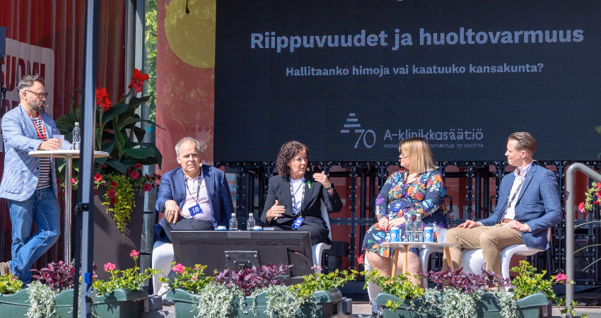 Paneelikeskustelu käynnissä SuomiAreena-tapahtumassa ulkolavalla kesäisessä säässä. Vasemmalla juontaja seisoo pöydän ääressä, oikealla neljä paneelikeskustelijaa istuu lavalla. Taustalla suuri näyttö, jossa lukee "Riippuvuudet ja huoltovarmuus – Hallitaanko himoja vai kaatuuko kansakunta?" sekä A-klinikkasäätiön 70-vuotisjuhlalogo. Lavalla kukkaistutuksia ja yleisölle suunnattuja istumapaikkoja. Tilanne vaikuttaa aktiiviselta ja keskustelevalta.