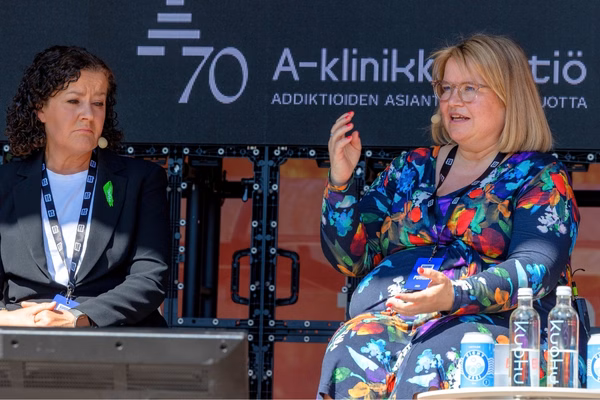 Kaksi henkilöä istuu lavalla SuomiAreena-tapahtumassa A-klinikkasäätiön järjestämässä keskustelussa. Vasemmalla oleva nainen kuuntelee vakavana, oikealla oleva nainen puhuu ja elehtii kädellään. Taustalla näkyy A-klinikkasäätiön 70-vuotisjuhlalogo ja teksti "Addiktioiden asiantuntijuutta 70 vuotta". Pöydällä on vesi- ja virvoitusjuomapulloja. Tapahtuma käynnissä aurinkoisessa ulkotilassa.