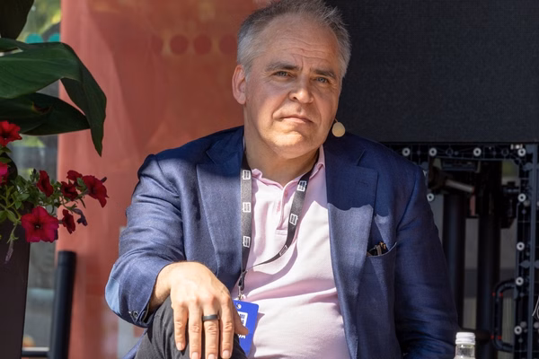 Henkilö istuu lavalla SuomiAreena-tapahtumassa ja katsoo vakavana kameraan. Hänellä on yllään sininen pikkutakki ja vaaleanpunainen kauluspaita, ja kaulassaan tapahtuman tunniste. Mies käyttää puhelinkuulokemikrofonia ja hänen vierellään on punaisia kukkia. Taustalla näkyy osittain musta näyttö ja tapahtumatekniikkaa. Aurinko paistaa kirkkaasti kasvoille.