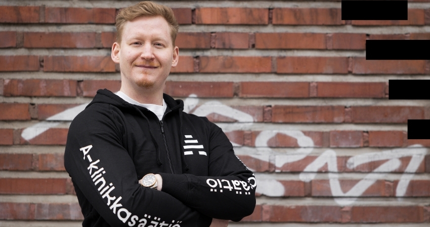 Kuvassa punahiuksinen henkilö seisoo tiiliseinän edessä ja katsoo lempeästi kameraan. Tiiliseinässä on graffiti taustalla