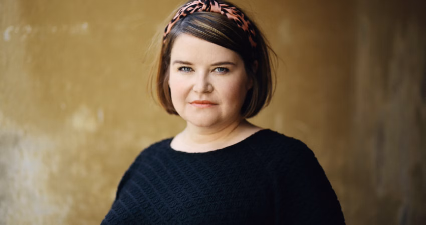 Kirjailija Anna Karhunen katsoo kameraan rauhallisesti ja suoraan. Hänellä on musta kuvioitu pusero ja vaaleanpunasävyinen leopardikuosinen hiuspanta. Taustalla on lämminsävyinen, rosoinen seinä. Karhunen on mukana A-klinikkasäätiön SuomiAreena-paneelikeskustelussa.