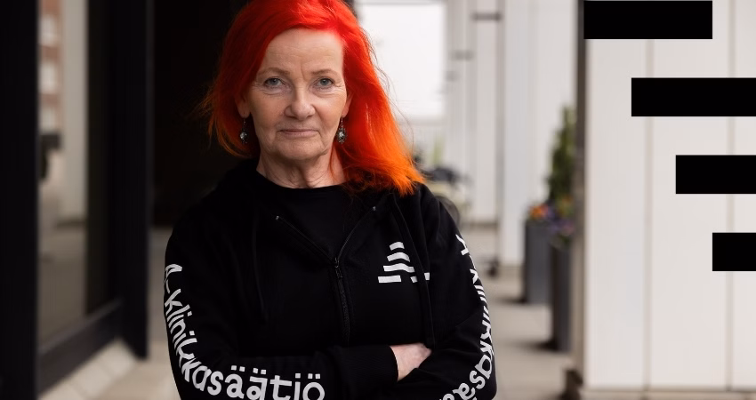 Punatukkainen henkilö seisoo kädet ristissä ulkotilassa, yllään musta huppari, jossa lukee "A-klinikkasäätiö". Hän katsoo suoraan kameraan itsevarmasti. Taustalla on vaalea rakennus ja ruukkukasveja. Kuvan oikeassa reunassa on mustia graafisia elementtejä.