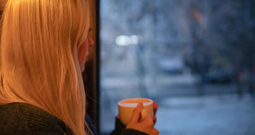 Nainen katsoo ikkunasta ulos kahvikuppi kädessä.